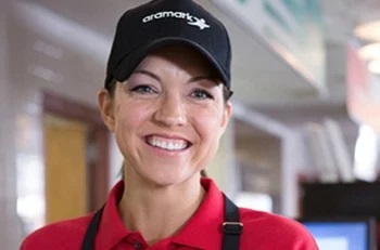 Smiling Aramark worker in an apron and a black Aramark hat.